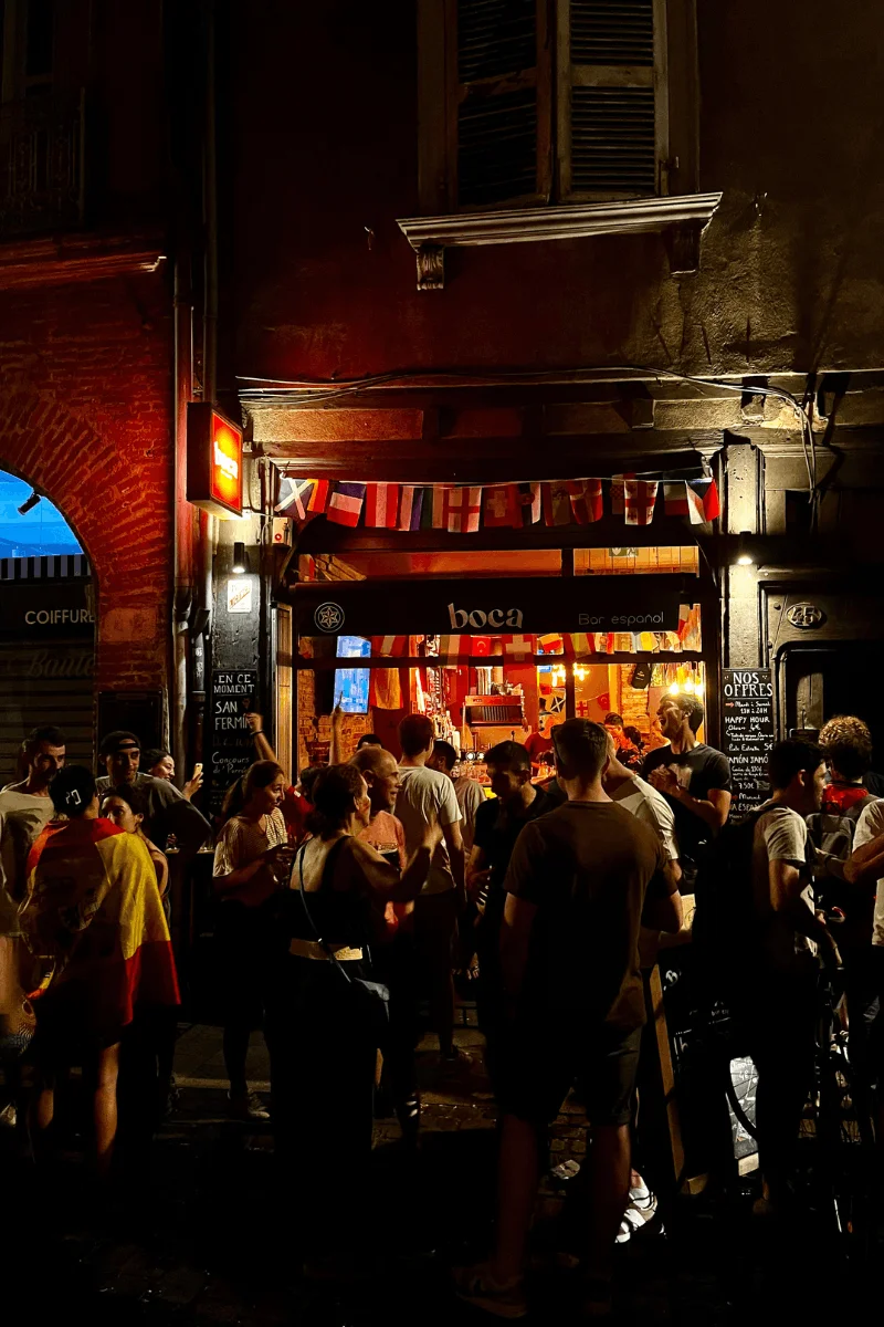 Foule de supporters devant le bar espagnol BOCA à Toulouse pour la finale de l'Euro 2024, avec une cliente drapée dans un drapeau espagnol, ambiance festive et conviviale pour les matchs de foot.
