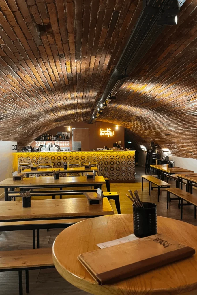 Vue d'ensemble de la cave du bar espagnol BOCA à Toulouse : table haute pour duo/trio, tablées à partager en bois massif, voûte en briques apparentes et lumière chaleureuse, créant une ambiance intimiste et authentique pour déguster des tapas ibériques.